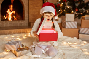 Extremely excited surprised little girl wearing white sweater and santa claus hat, opens gift box with something glowing, sitting on floor near Christmas tree, present boxes and fireplace.